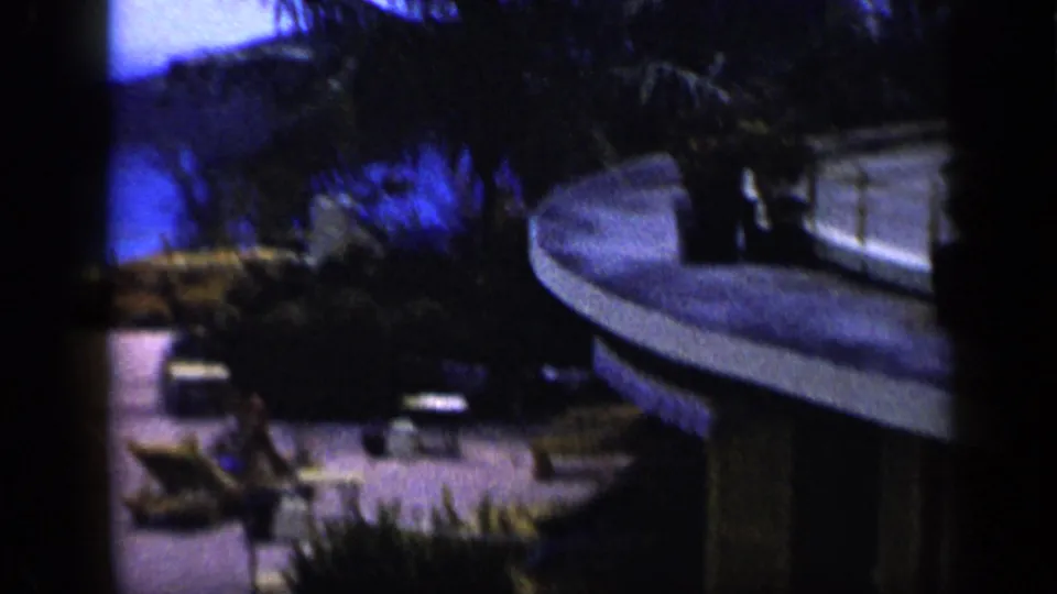 Grainy 1959 Venezuela resort view with lounge chairs, palm vegetation, terrace paving, and part of a hotel structure.