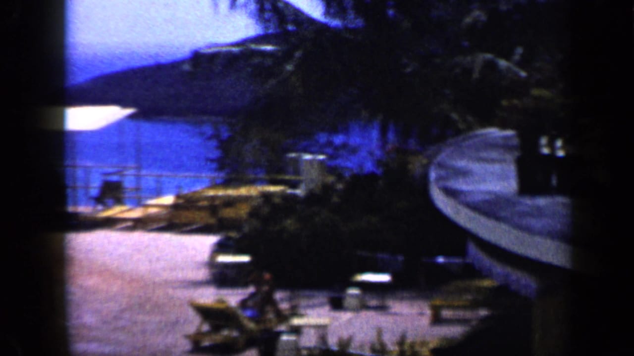 Coastal resort terrace in Venezuela with lounge chairs, palms, blue sea, and hills visible in 1959 home-movie footage.