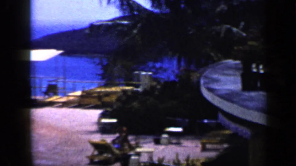 Coastal resort terrace in Venezuela with lounge chairs, palms, blue sea, and hills visible in 1959 home-movie footage.