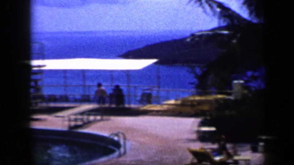 1959 Venezuela home-movie frame showing a resort pool deck, white cabana shade, guests and blue coastal water beyond.