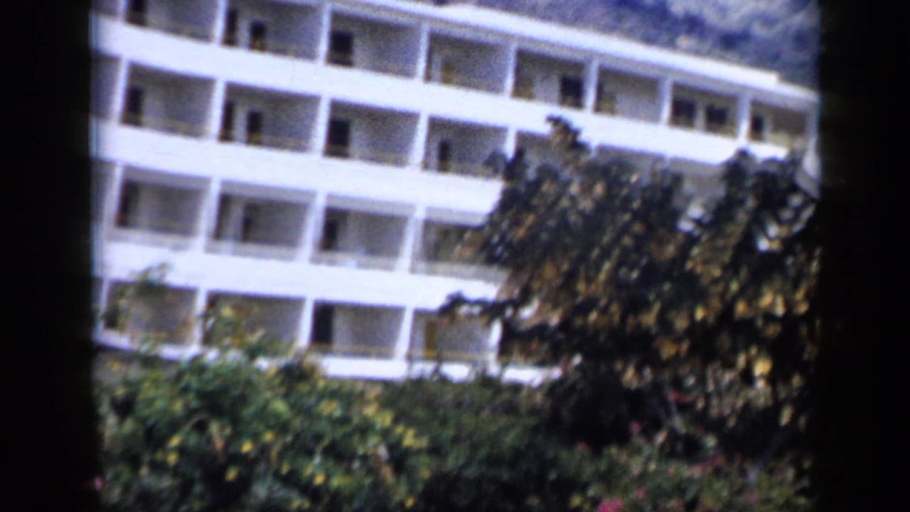A white multi-story hotel facade rises behind lush greenery and hillside plants in 1959 Venezuela travel footage.