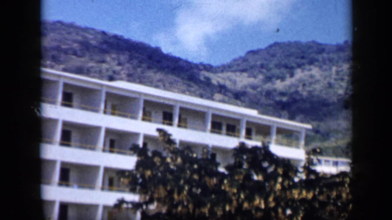 1959 Venezuela home-movie frame showing a white balcony-lined building partly hidden by trees below rugged hills.