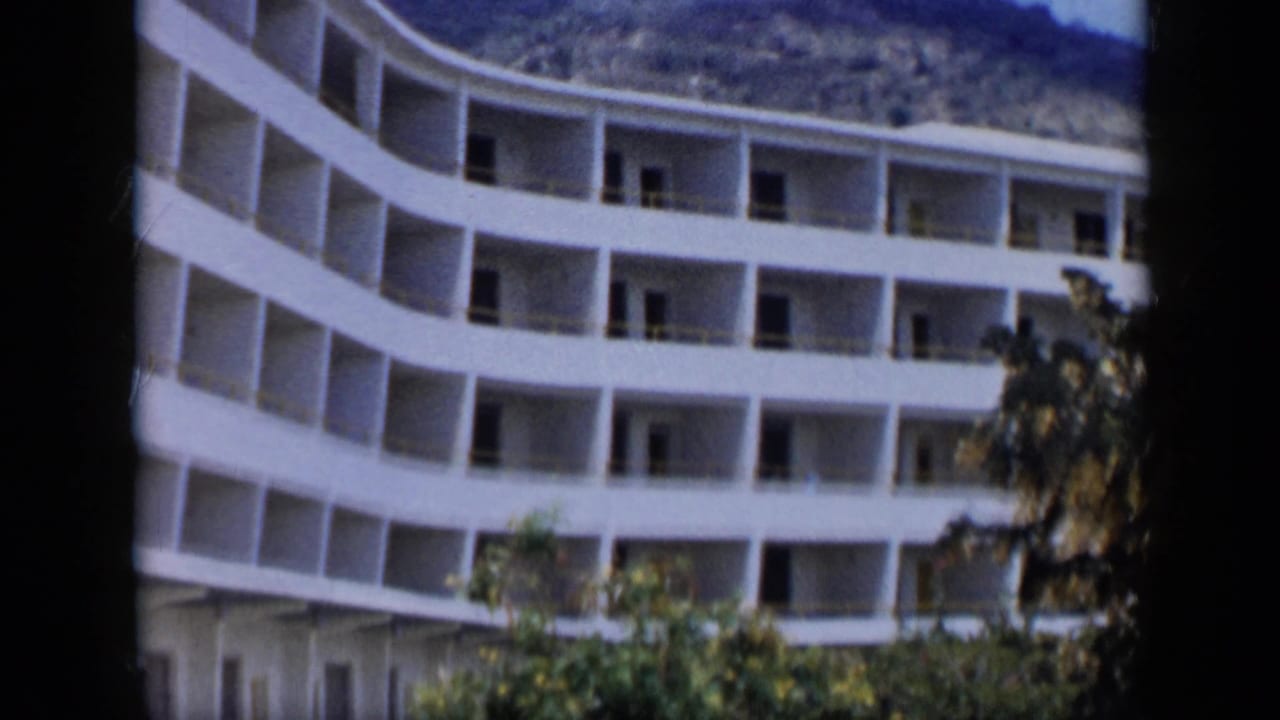 1959 Venezuela home-movie frame of a large curved white building with balcony rows, trees in front, and hills behind.