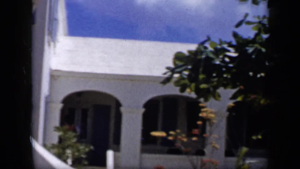 1959 Venezuela 8mm frame of a white building with arched openings behind orange tree foliage and shadowed foreground trunks.
