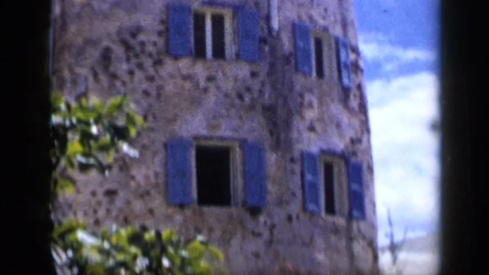 1959 Venezuela 8mm frame of a sunlit stone facade with multiple blue shutters and foliage along the lower edge.