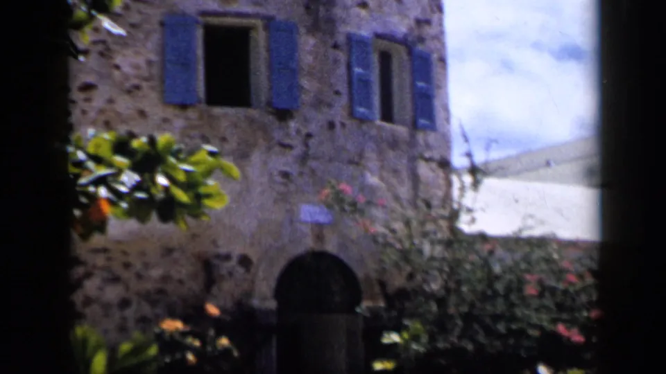 1959 Venezuela 8mm frame showing a stone exterior with blue shutters, an arched doorway, flowering shrubs, and a white adjacent wall.