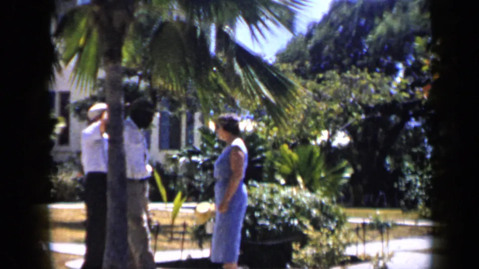 In a 1959 Venezuela garden scene, a man in light clothing and a woman in a blue dress stand near palms and shrubs.