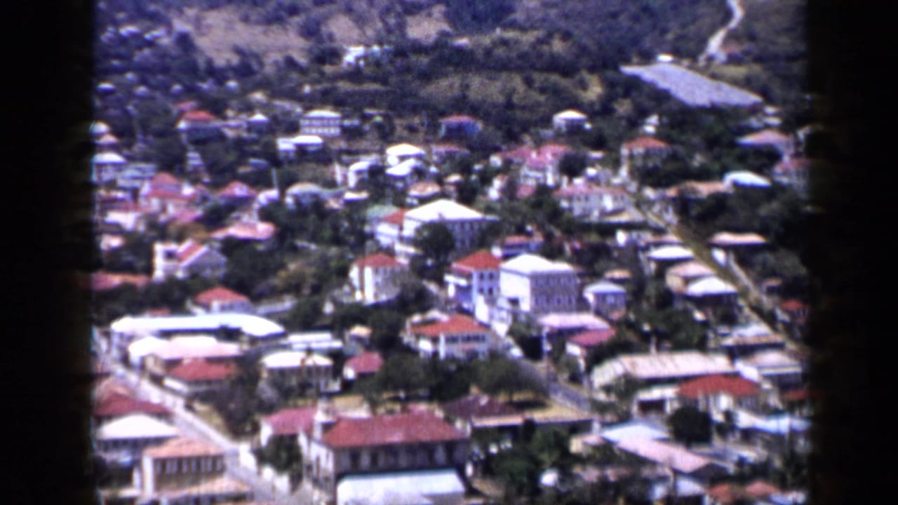 Hillside city with clustered buildings, red roofs, roads, and mountains beyond.