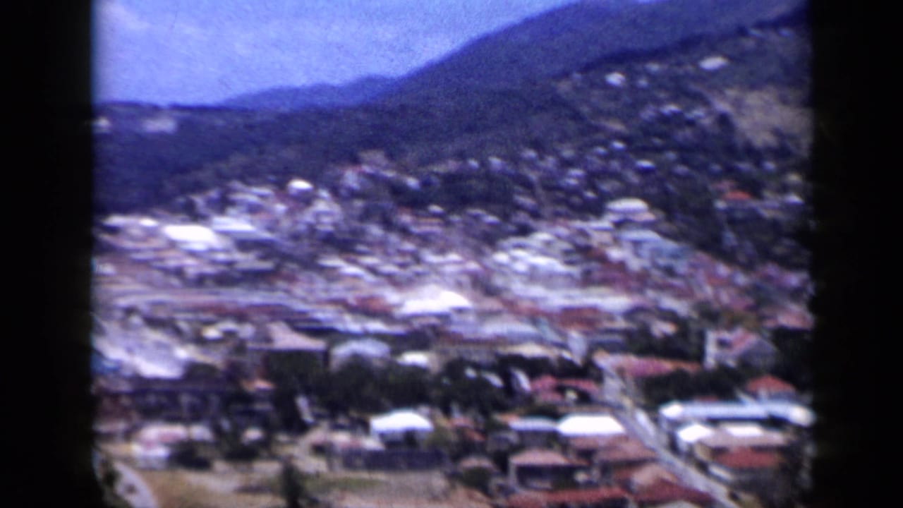 1959 Venezuela frame showing a soft aerial-style view over hillside houses, roads, and pale roofs, with mountains and coastline nearby.