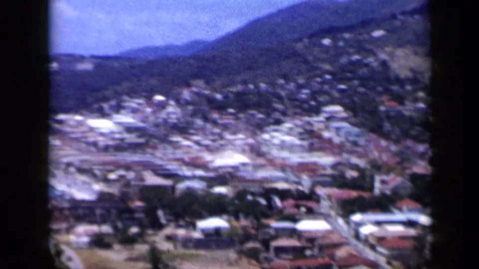 1959 Venezuela frame showing a soft aerial-style view over hillside houses, roads, and pale roofs, with mountains and coastline nearby.