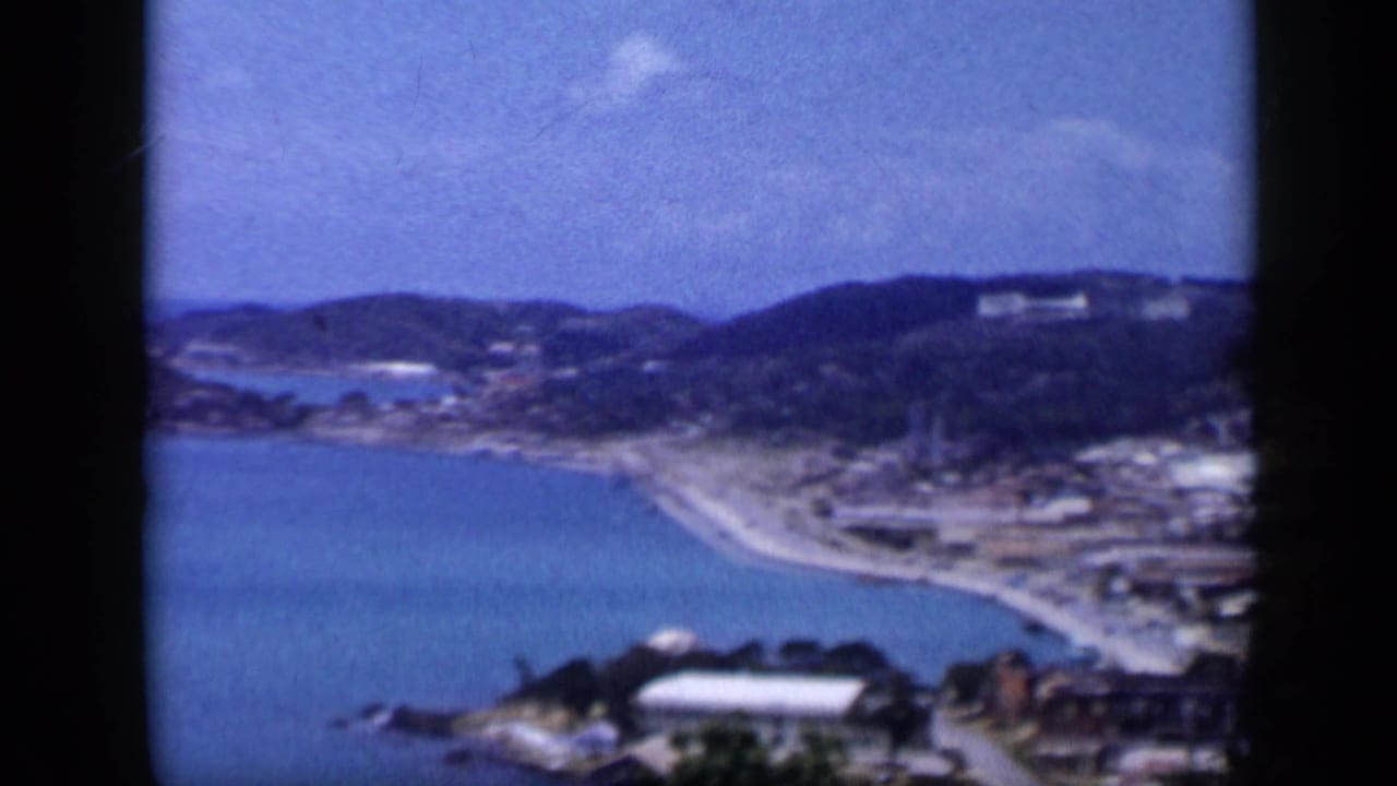 1959 Venezuela coastal view with a calm blue bay, curved shoreline road, buildings near the water, and dark hills beyond.