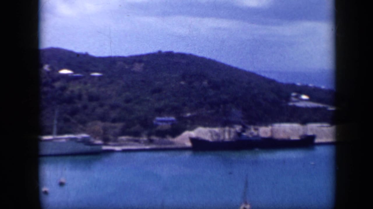 1959 Venezuela harbor view with calm blue water, docked boats, wooded hills, and soft 8mm film edges.