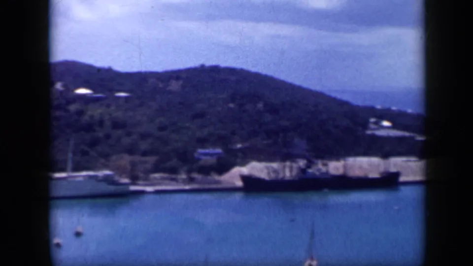 1959 Venezuela harbor view with calm blue water, docked boats, wooded hills, and soft 8mm film edges.
