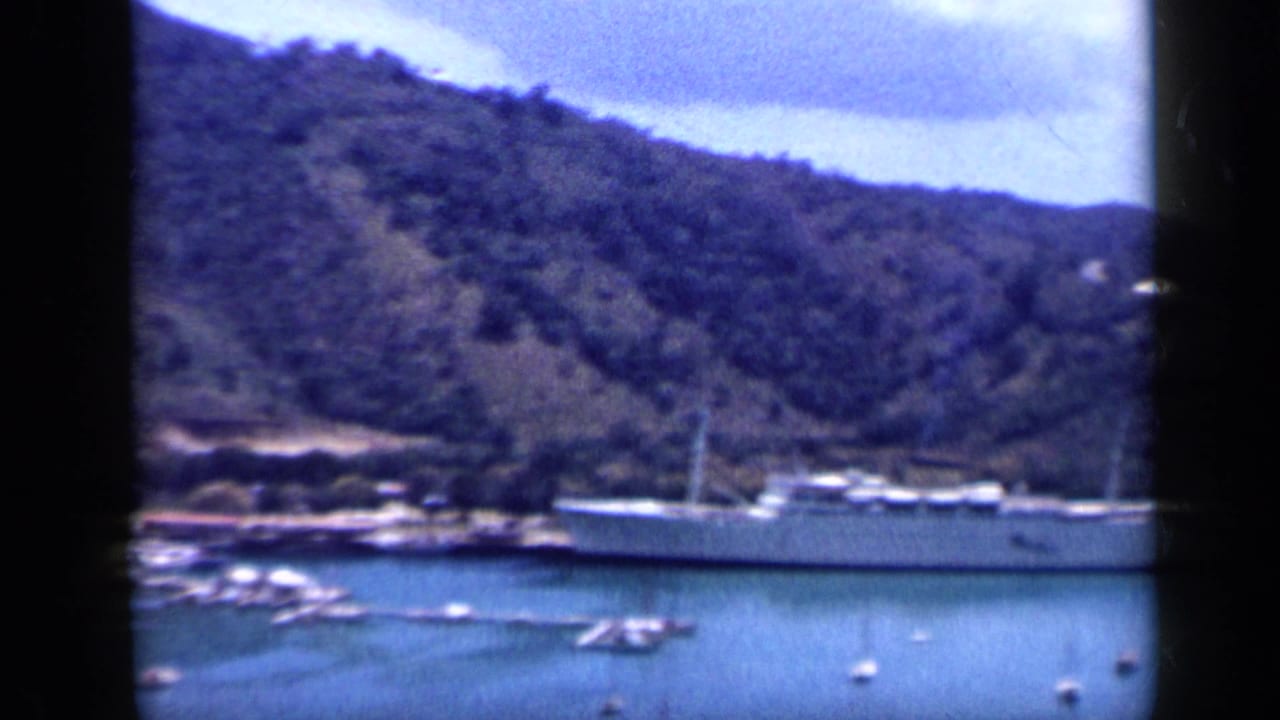 1959 harbor frame showing a pale moored ship, small boats on the water, and a wooded hillside in Venezuela.