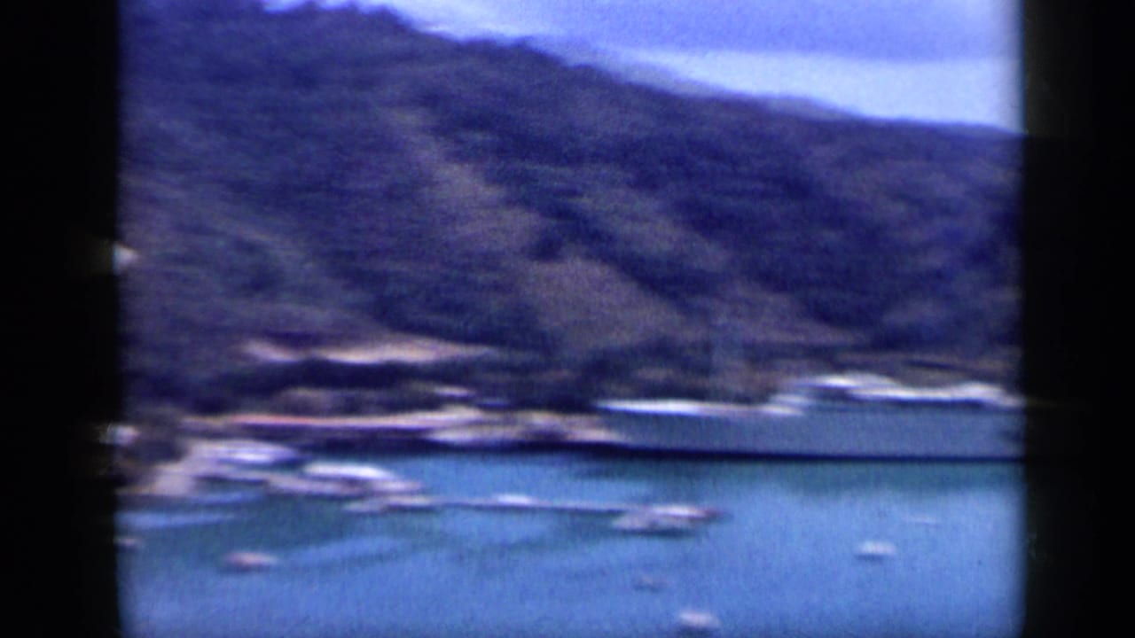 Wide 8mm view of a Venezuelan hillside harbor with a long pale vessel, small boats, and calm water.