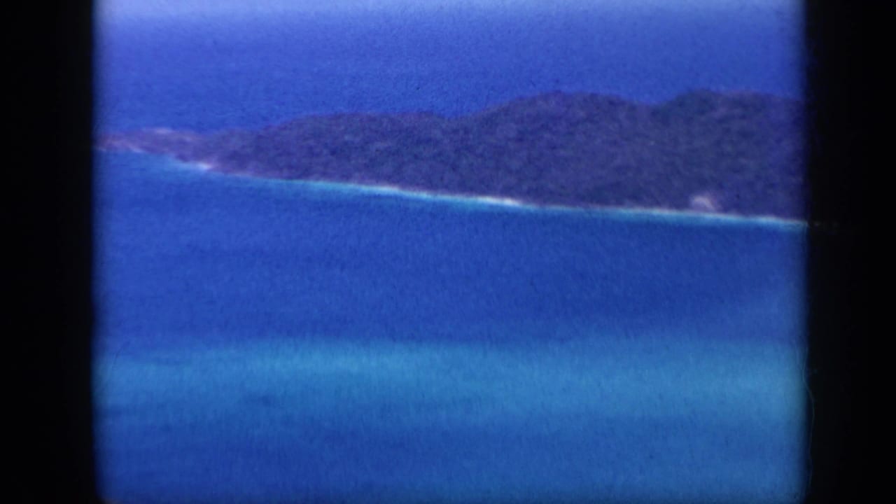 1959 Venezuela coastal view with a forested island ridge, bright blue sea, and a narrow light surf line along the shore.