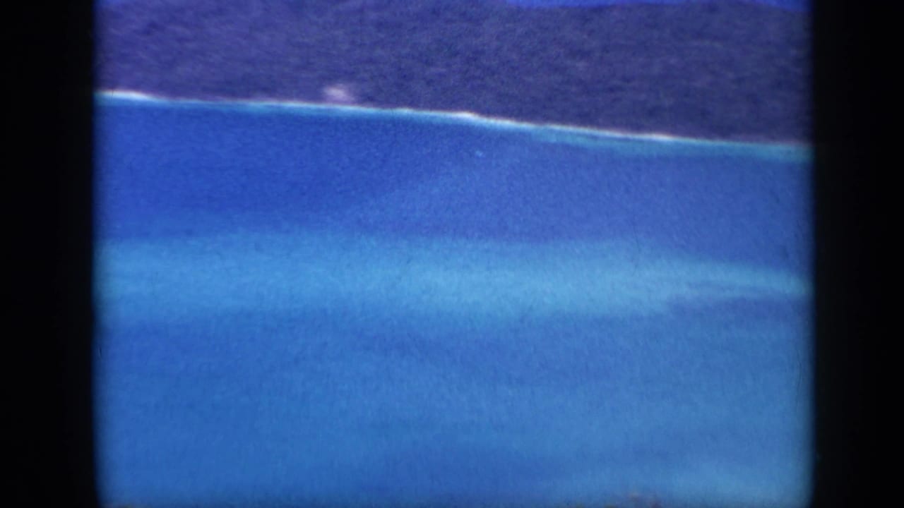1959 Venezuela frame showing a closer section of tropical shoreline with forested land, pale surf, and layered blue shallows.
