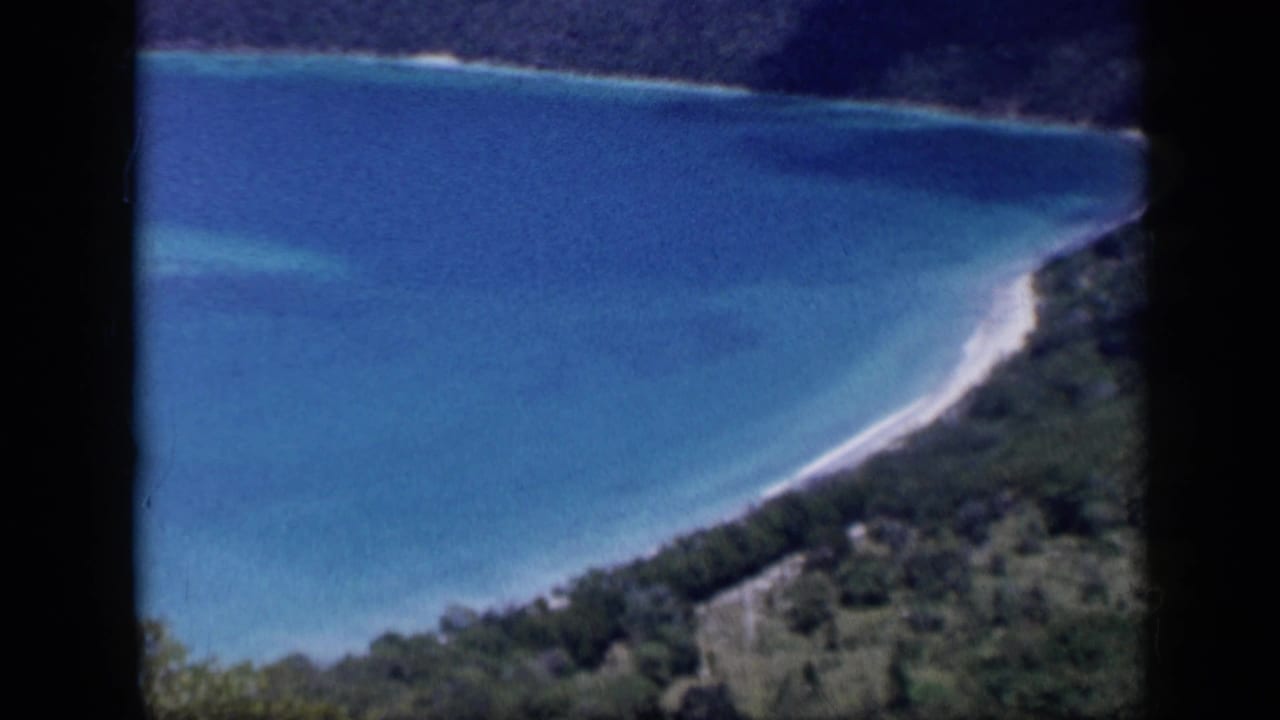 1959 Venezuela 8mm frame showing a turquoise coastal bay, white surf line, beach, and dense green hillside from an elevated view.