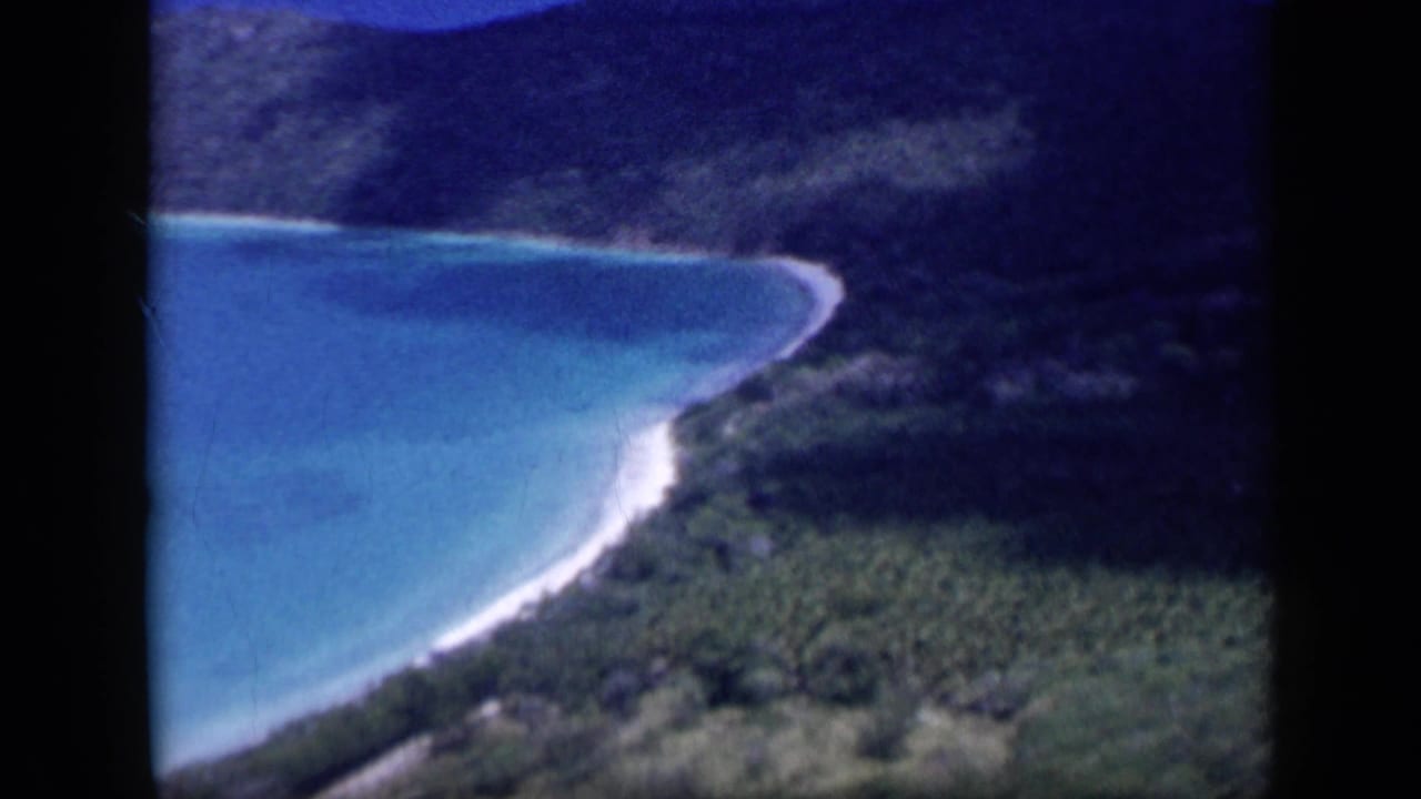 1959 Venezuela 8mm frame of a blue-green bay and pale shoreline bordered by wooded hills, seen from above with visible film gate edges.