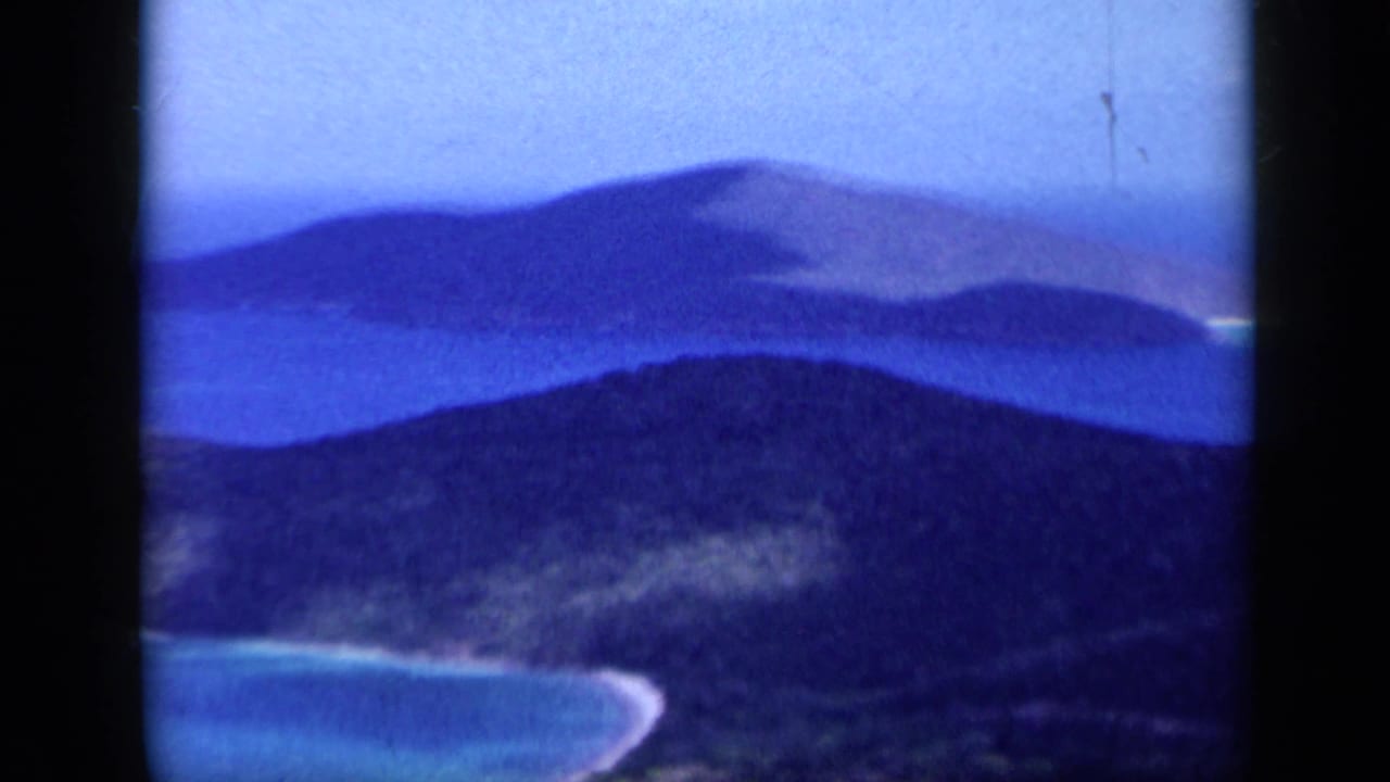 1959 Venezuela 8mm frame looking across layered coastal hills, blue water, and a pale beach curve in soft, saturated color.
