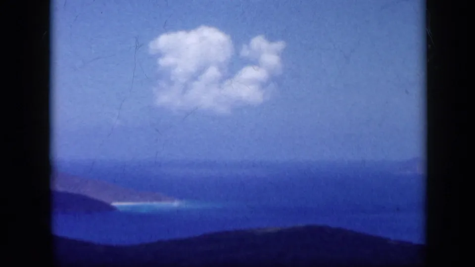 1959 Venezuela 8mm frame showing blue sea, distant land, dark hills, and white clouds from an elevated viewpoint.