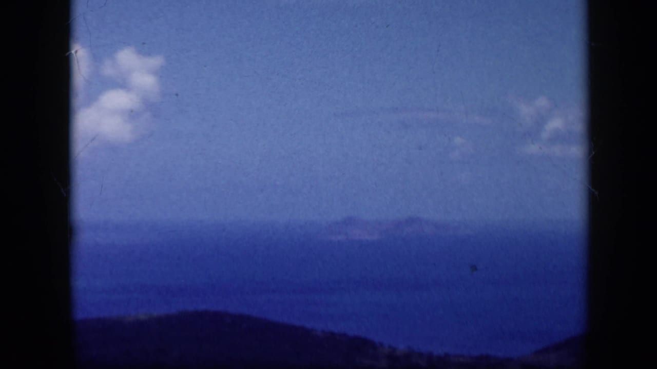 1959 Venezuela 8mm frame with distant island forms across blue water, a dark hillside foreground, and scattered clouds.