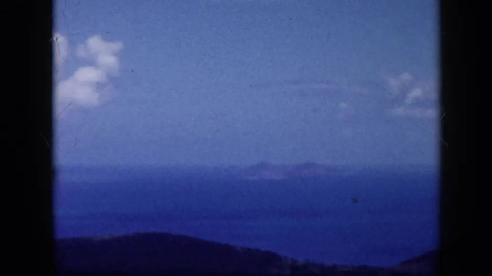 1959 Venezuela 8mm frame with distant island forms across blue water, a dark hillside foreground, and scattered clouds.