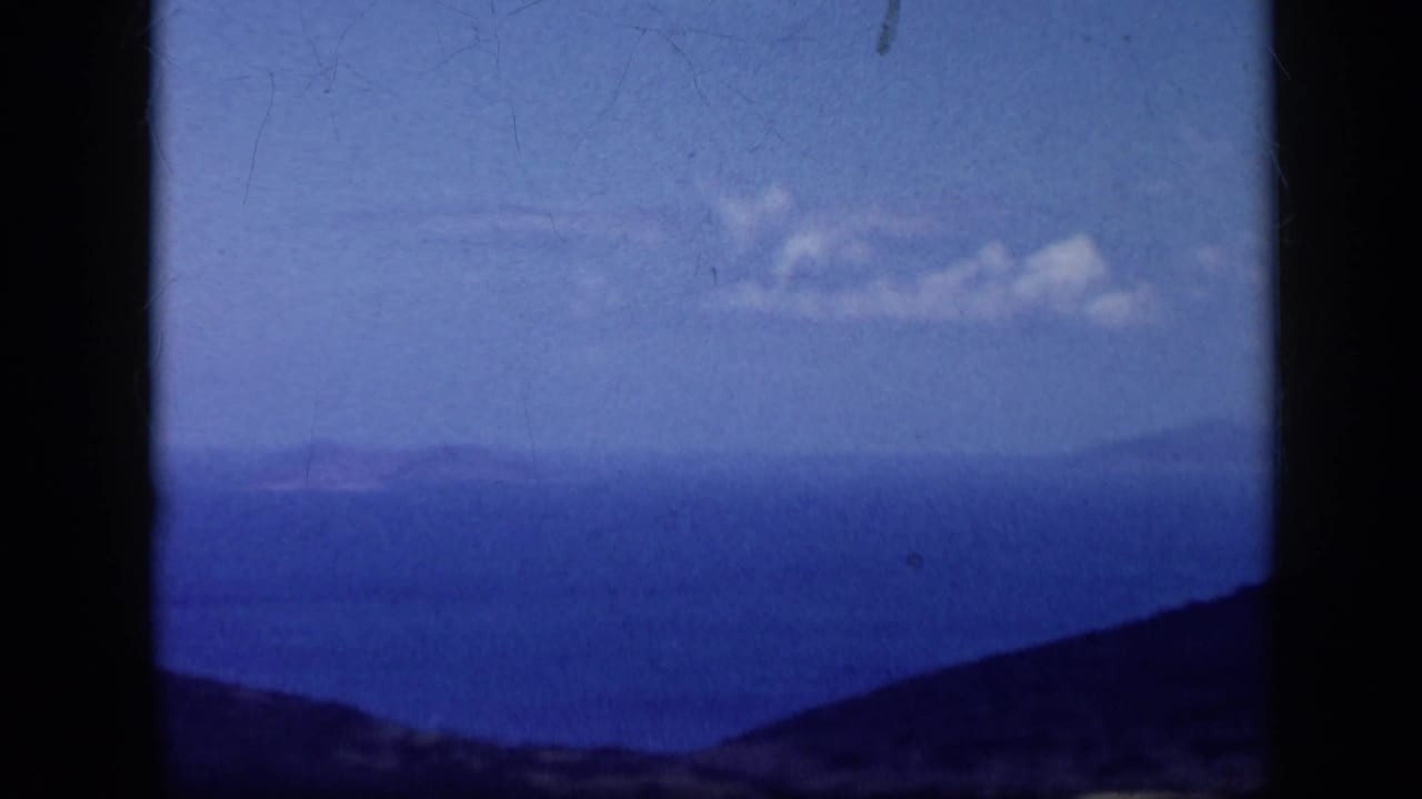 1959 Venezuela 8mm frame showing a hazy ocean panorama, rolling dark hills, and small clouds in a bright sky.