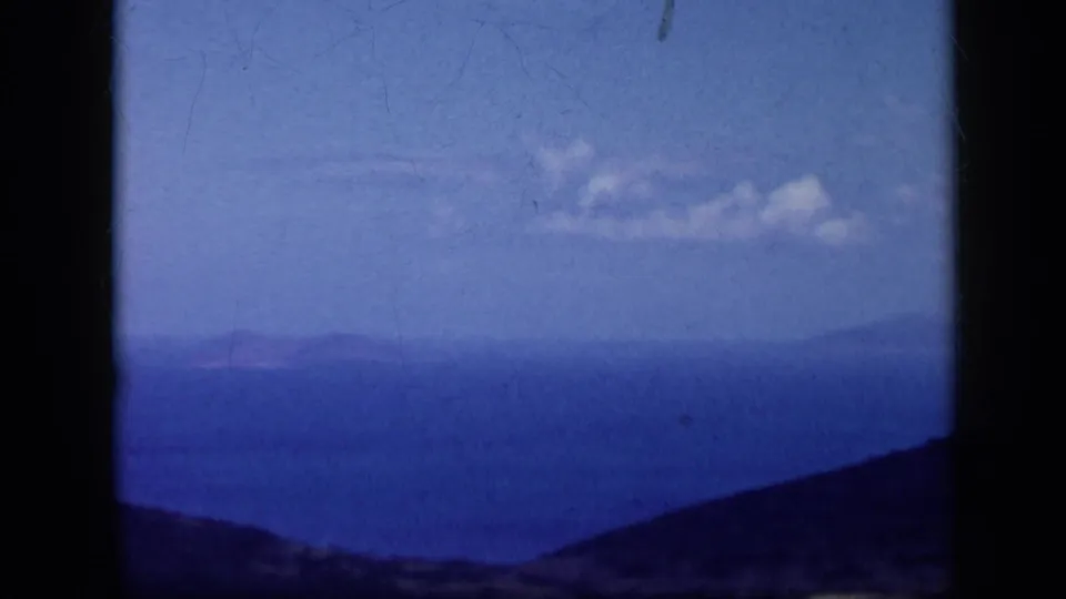 1959 Venezuela 8mm frame showing a hazy ocean panorama, rolling dark hills, and small clouds in a bright sky.