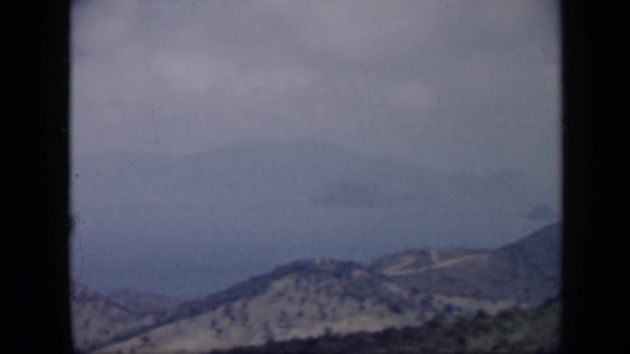 1959 Venezuela 8mm frame showing brushy mountain ridges descending toward hazy coastal water beneath clouds.