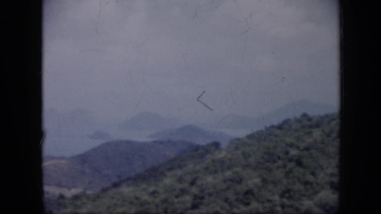 1959 Venezuela 8mm frame with a foreground hillside, layered mountains, and pale blue water in the distance.