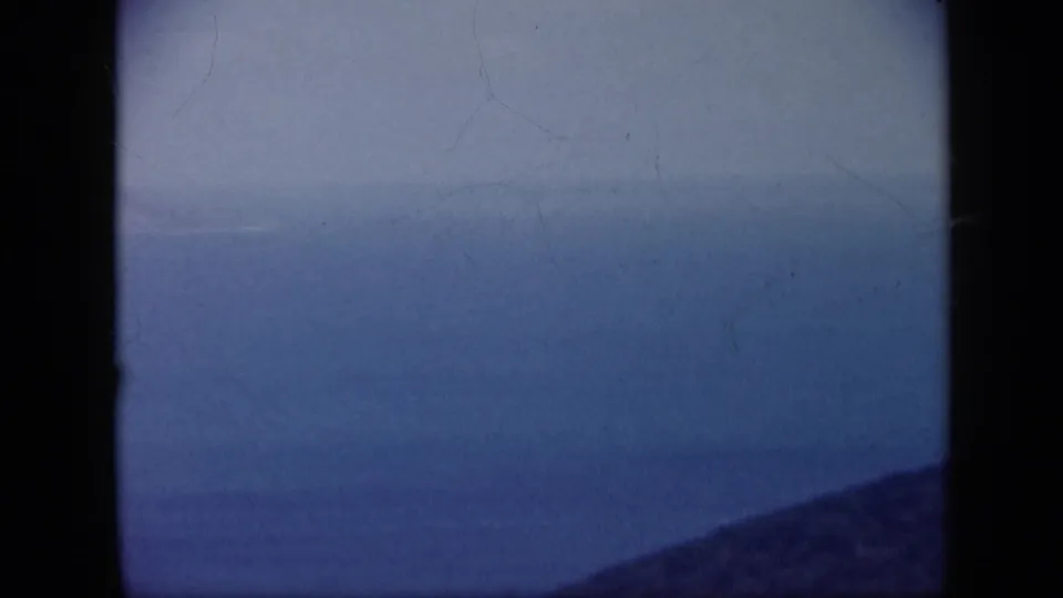 1959 Venezuela 8mm frame looking across blue coastal water toward a faint shoreline, with a dark ridge at the bottom edge.