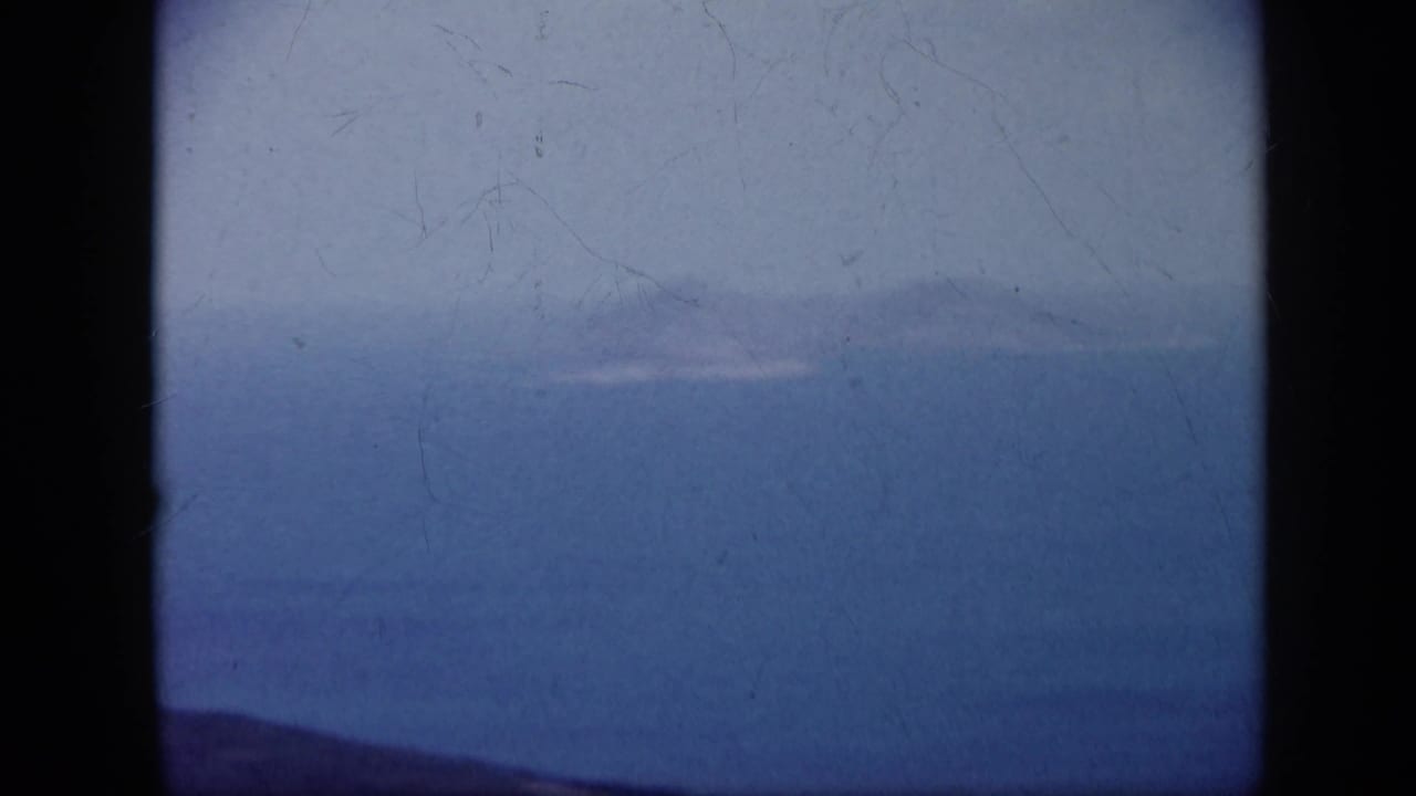 1959 Venezuela 8mm frame showing a broad ocean inlet, distant hazy landforms, and a dark ridge along the lower edge.