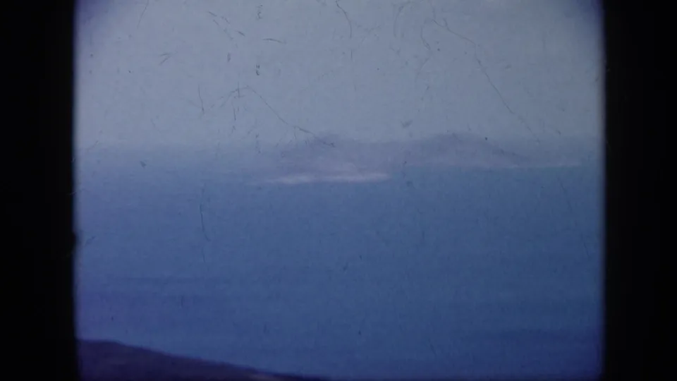 1959 Venezuela 8mm frame showing a broad ocean inlet, distant hazy landforms, and a dark ridge along the lower edge.
