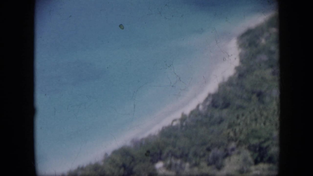 1959 Venezuela coastal frame with a bright beach line, turquoise sea, and thick greenery seen from an elevated viewpoint.