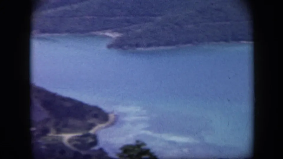 1959 Venezuela home-movie frame showing blue water, dark hills, and a curved shoreline seen through an airplane window.
