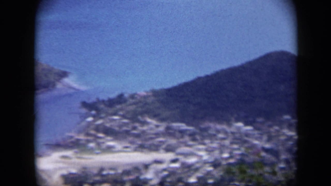 1959 Venezuela aerial view with a clustered shoreline town, pale beach, blue water, and dark mountain slopes.