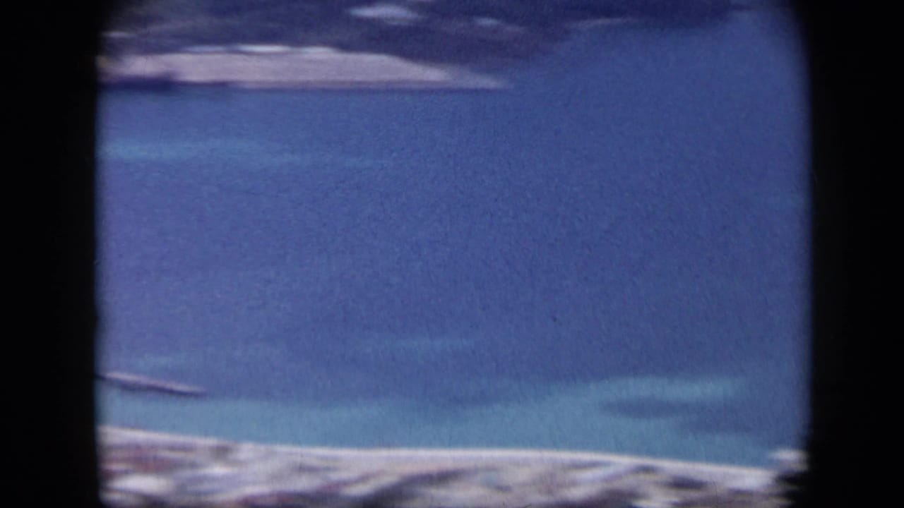1959 Venezuela aerial frame showing a blue bay, pale beach, and distant shoreline through soft 8mm film texture.