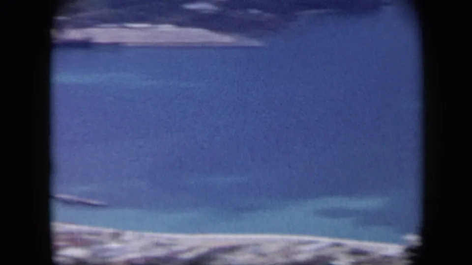 1959 Venezuela aerial frame showing a blue bay, pale beach, and distant shoreline through soft 8mm film texture.