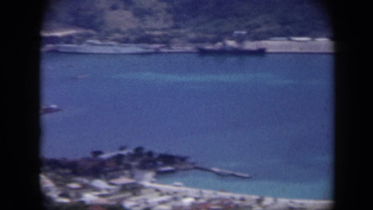 1959 Venezuela aerial 8mm view over a coastal harbor with turquoise water, shoreline buildings, and a ship offshore.