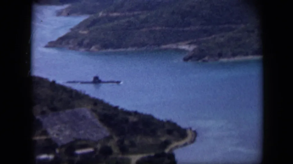 1959 Venezuela aerial frame with coastal water, green shoreline, a road, and a low vessel or submarine-like craft offshore.