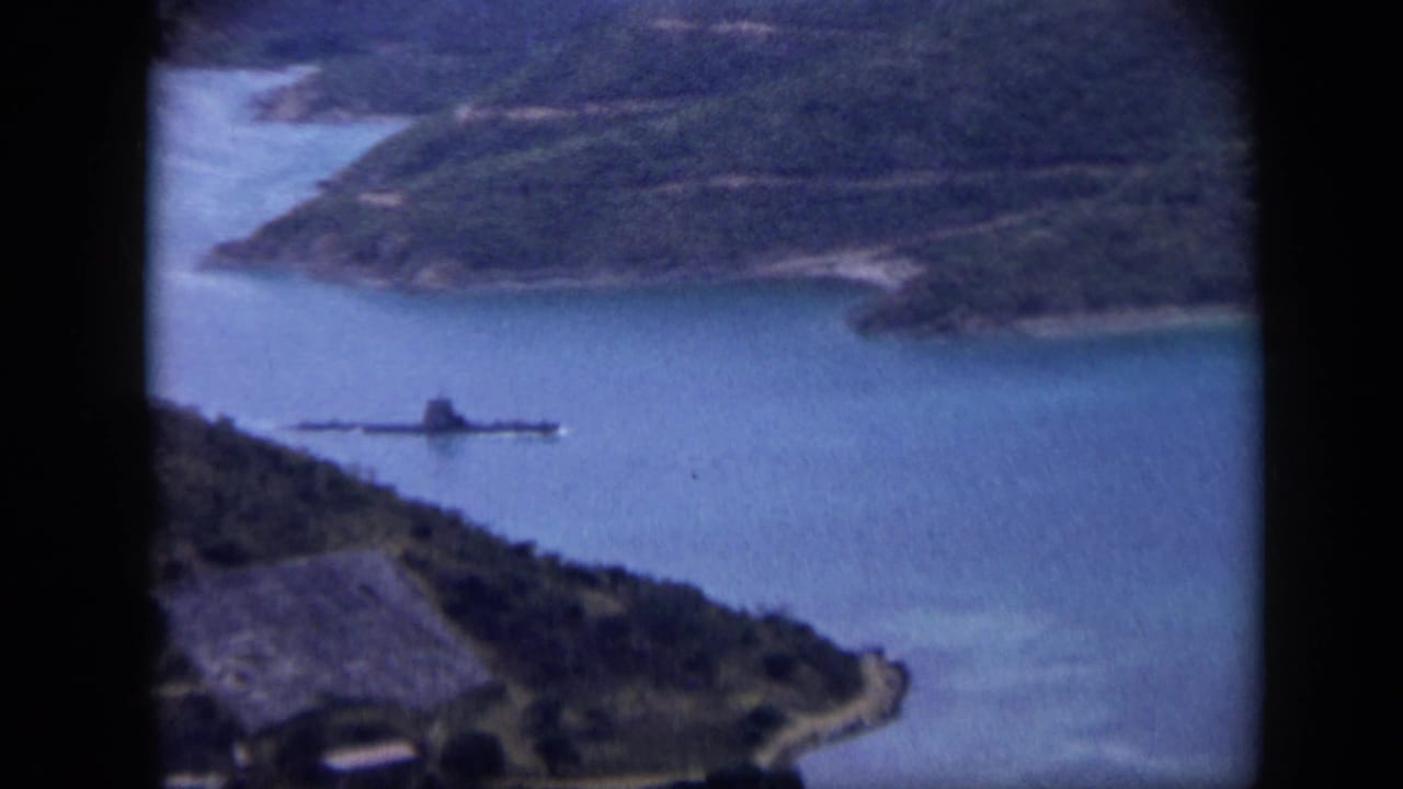 Long dark vessel crossing blue harbor water below green hills.