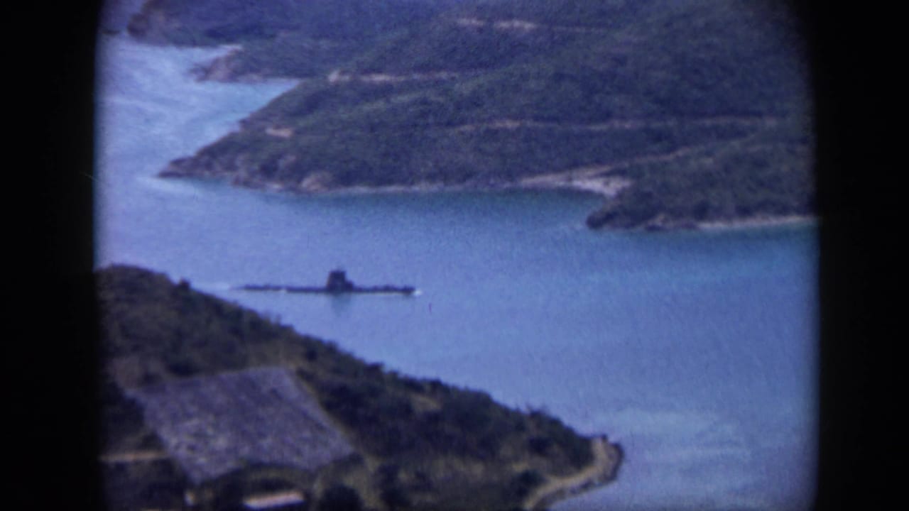 Elevated 1959 Venezuela view of a long dark harbor vessel against blue water and wooded coastal hills.