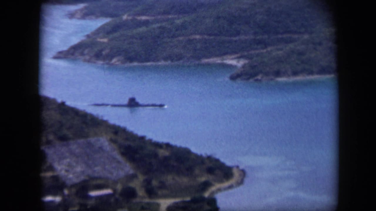 Home-movie harbor view from Venezuela in 1959, with a dark vessel crossing calm water below a green hillside.