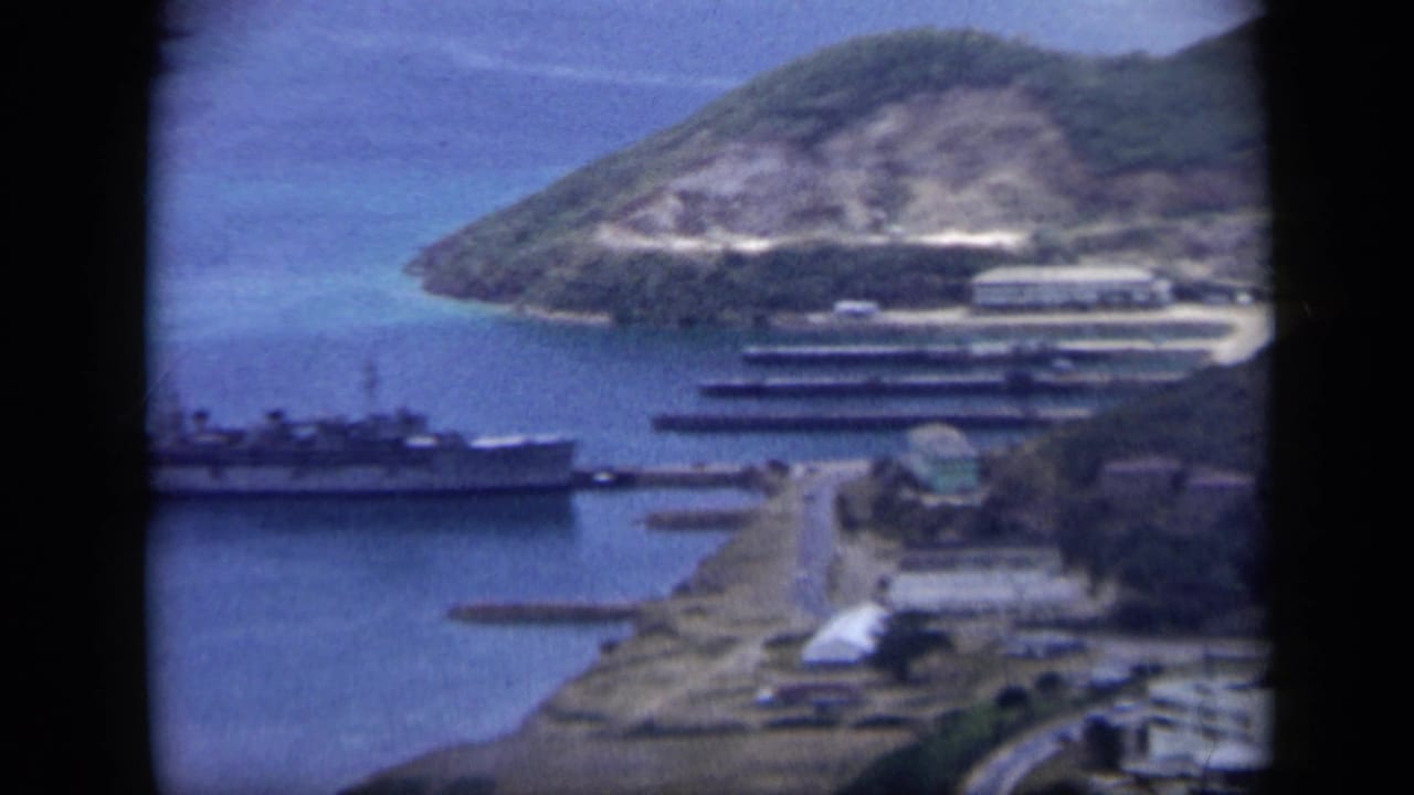 1959 Venezuela harbor frame showing docks, moored vessels, waterfront buildings, and a hillside road beside blue water.