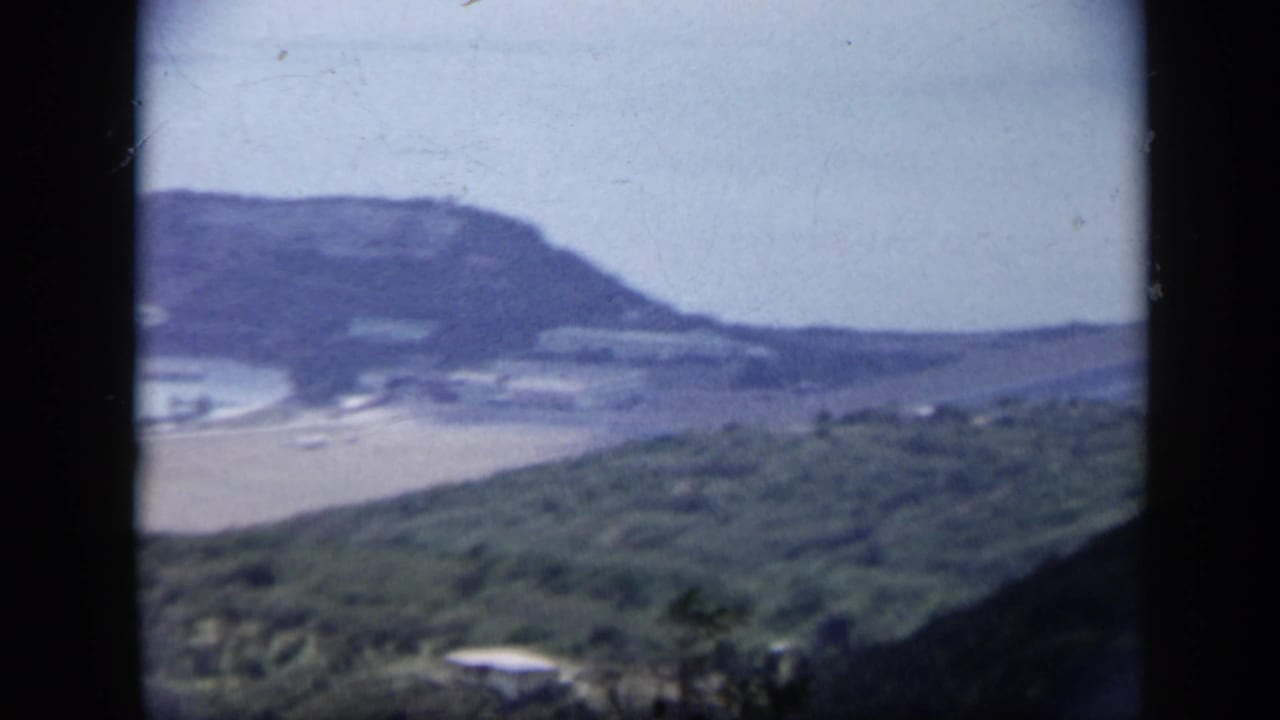 1959 Venezuela 8mm frame from an elevated vantage point, looking over a sandy shore, bay water, green hillside, and distant headland.