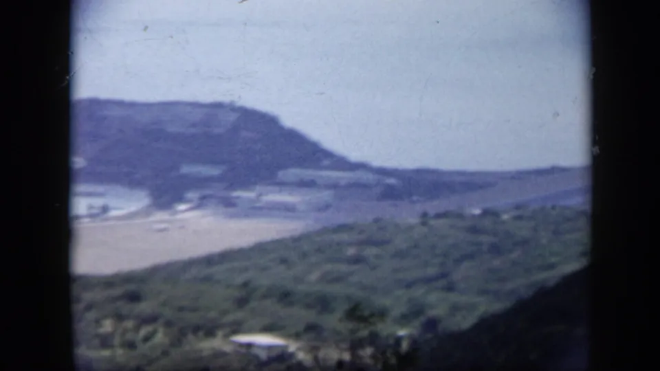 1959 Venezuela 8mm frame from an elevated vantage point, looking over a sandy shore, bay water, green hillside, and distant headland.