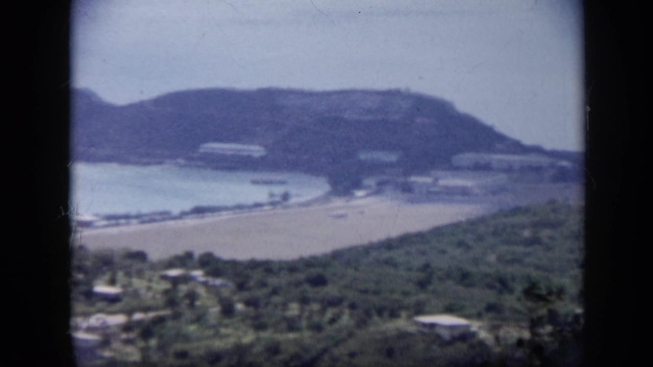 1959 Venezuela 8mm frame showing an elevated look toward a pale beach, quiet bay, green hills, and scattered shoreline structures.