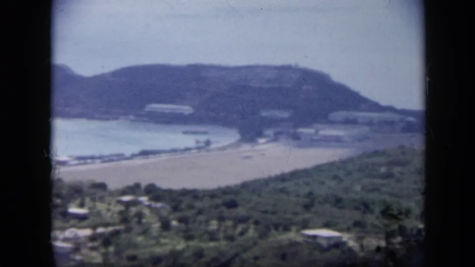 1959 Venezuela 8mm frame showing an elevated look toward a pale beach, quiet bay, green hills, and scattered shoreline structures.