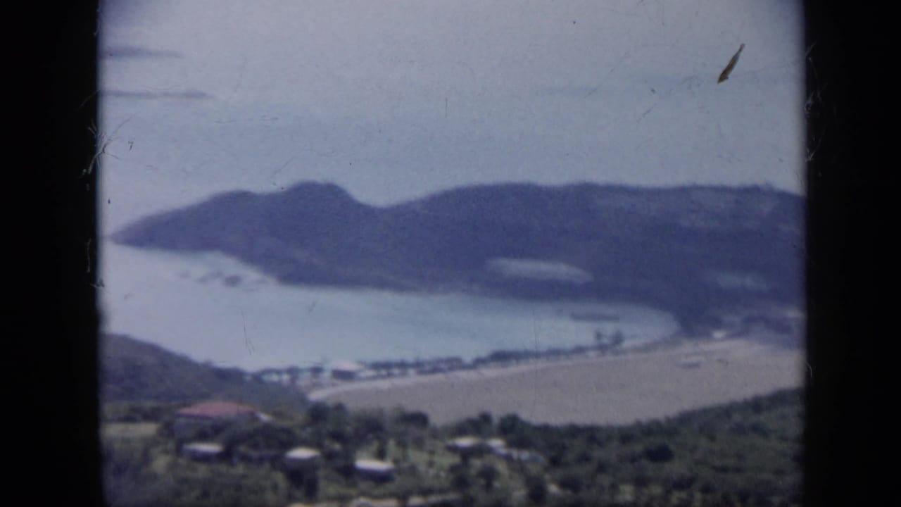 1959 Venezuela 8mm frame with a wide elevated view of a sheltered bay, sandy shoreline, dark wooded hills, and small distant buildings.